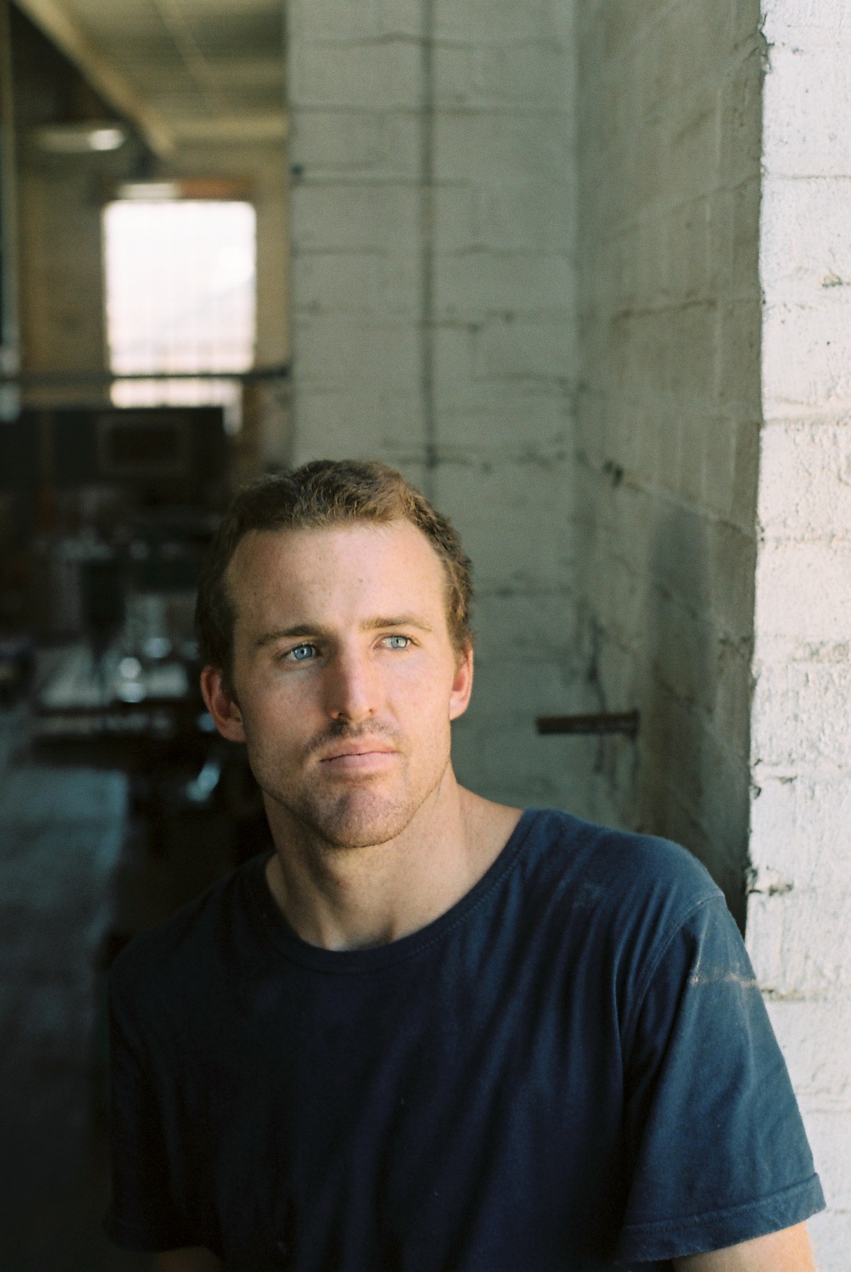 Portrait of Alex Fossilo, a designer-craftsman, in an industrial workshop with white brick walls.