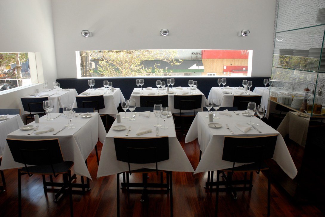 Modern restaurant dining room with white-clothed tables, black chairs, and large windows overlooking São Paulo.