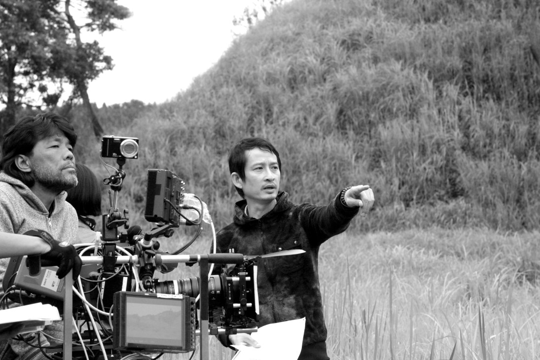 Director Trần Anh Hùng gestures while directing a film scene with cinematographer and camera equipment in a mountainous forest setting.