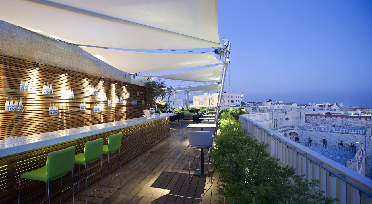Rooftop bar with wooden slat wall, green bar stools, retractable awning, and Jerusalem cityscape view