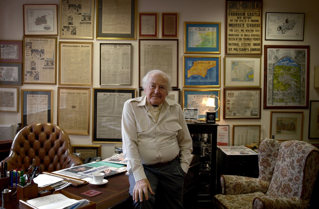 Sir Ray Tindle, an elderly man in a cream shirt, sits at his desk surrounded by framed historical newspapers and maps on the walls behind him.