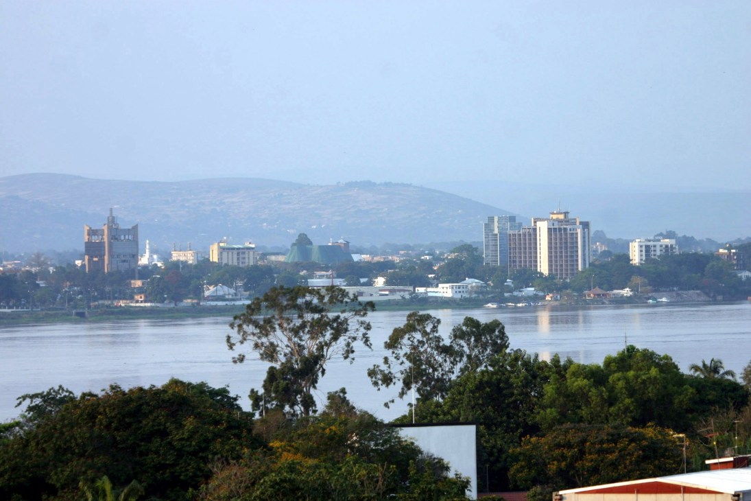 Brazzaville skyline with colonial architecture across the Congo River, with forested riverbank in foreground.