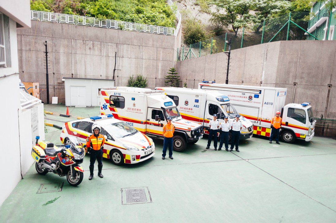 Hong Kong ambulance command staff stand with their fleet including motorcycles, cars and ambulances in a depot courtyard.