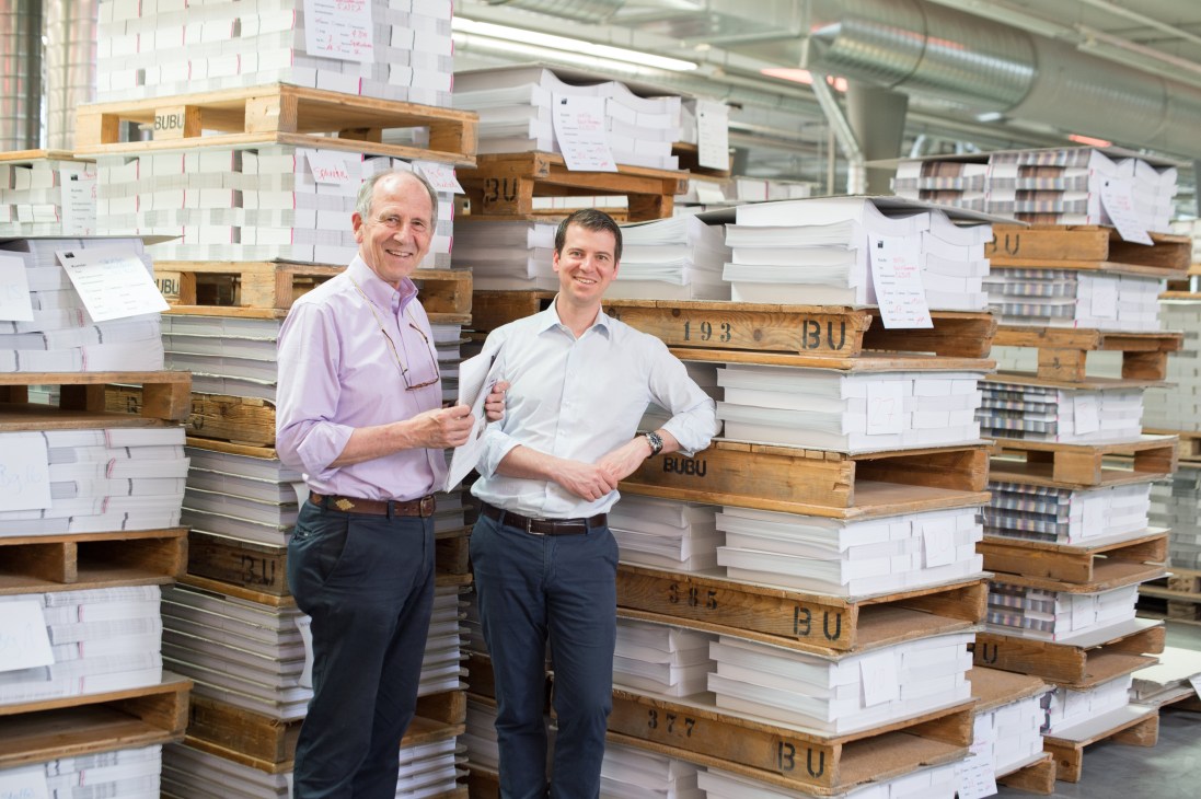 Two men stand smiling in a bookbinding facility surrounded by stacks of bound books on wooden pallets labeled BUBU.
