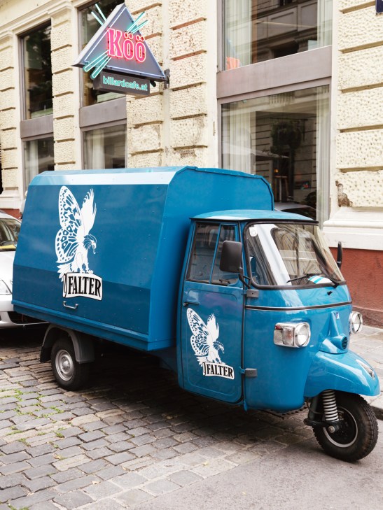 Blue three-wheeled delivery vehicle branded with Falter magazine logo and butterfly symbol on a Vienna street.
