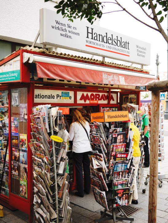 A red newsstand kiosk displaying magazines and newspapers with Handelsblatt signage visible above.