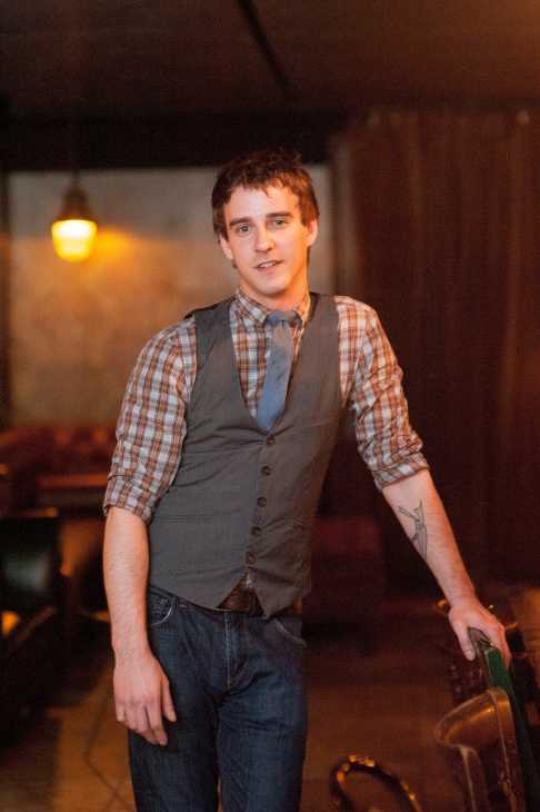 Man in gray vest and plaid shirt holding a bottle in a dimly lit bar setting.