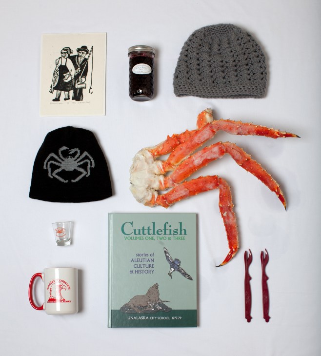 Flat lay of Unalaska souvenirs including block print, jam jar, knitted hat, coffee mug, red king crab, shot glass, Cuttlefish book, and crab forks.