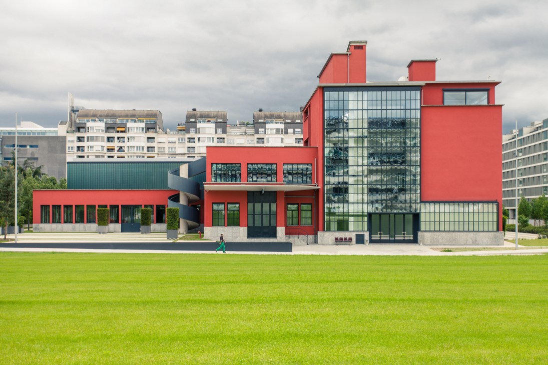 Red modernist building with large windows overlooking a manicured green lawn in an urban setting.