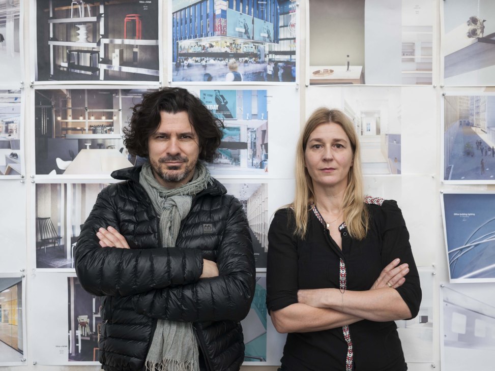 Architects Pierre Jorge Gonzalez and Judith Haase stand with crossed arms in front of architectural renderings and building photographs displayed on a white wall.