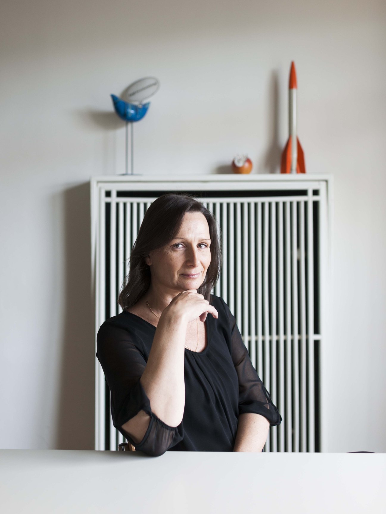 Portrait of Sabine Ofenbach, managing director of Texte zur Kunst magazine, posing thoughtfully with her hand near her face in front of a white radiator with vertical slats.