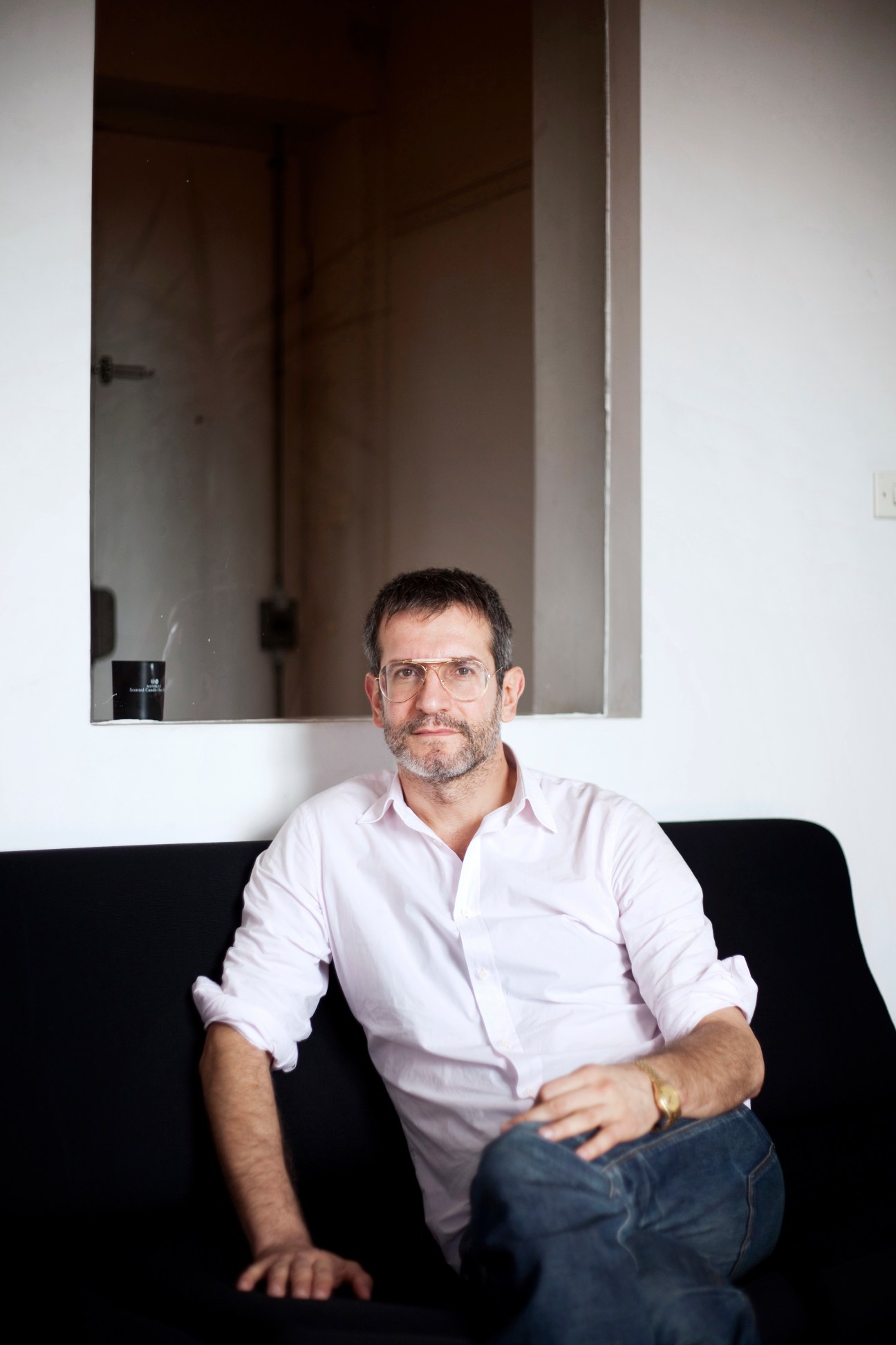 Man in pink shirt and glasses sitting on black sofa against white wall with dark window