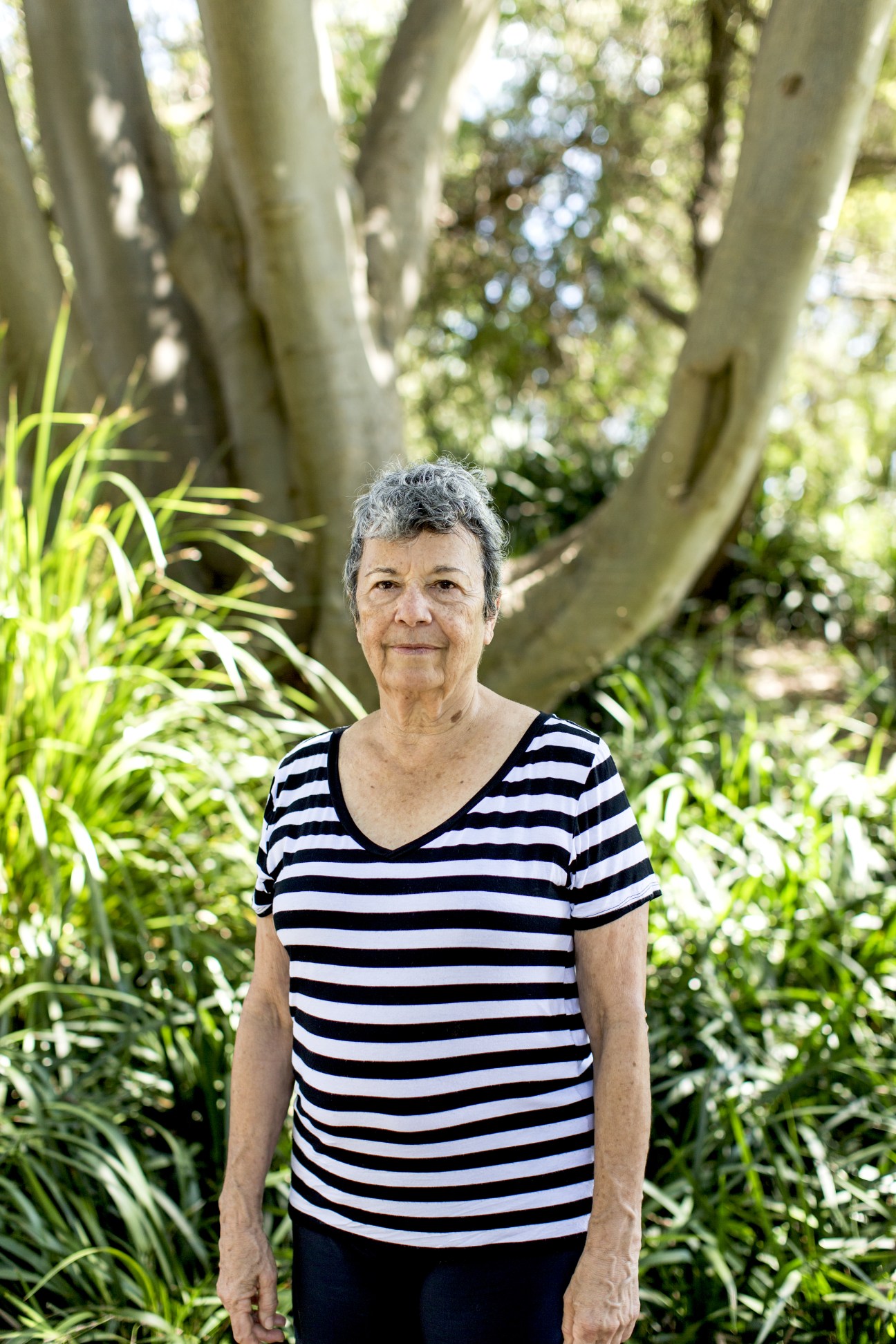 Pat O'Shane, barrister, standing in a garden wearing a striped shirt beneath arching tree branches.