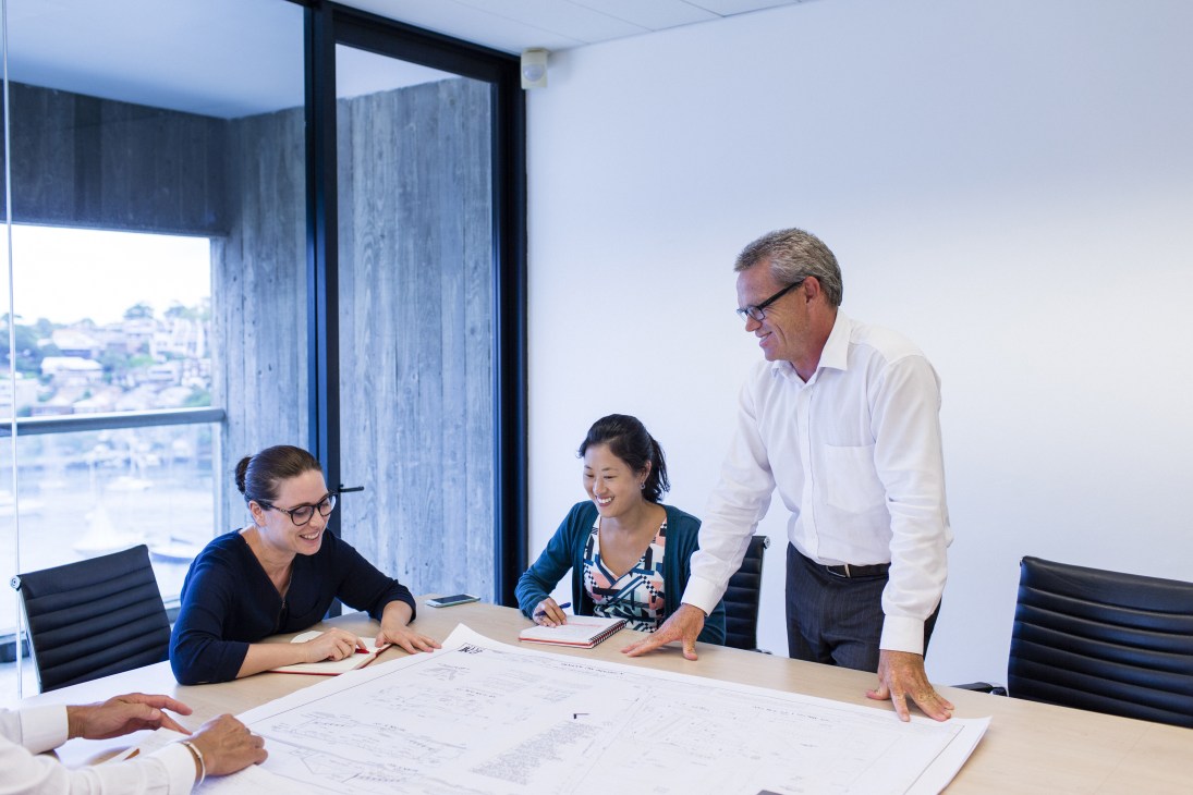 Three team members reviewing architectural plans at a table in a high-rise office with city views.