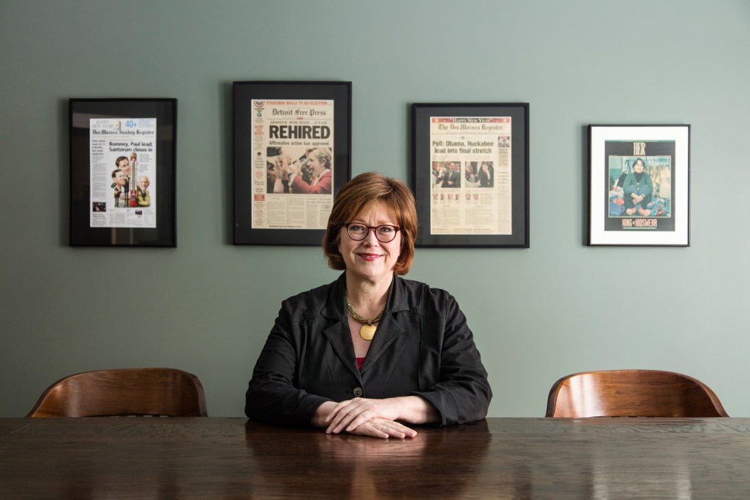 Ann Selzer seated at a desk with framed newspaper front pages displayed on the wall behind her.