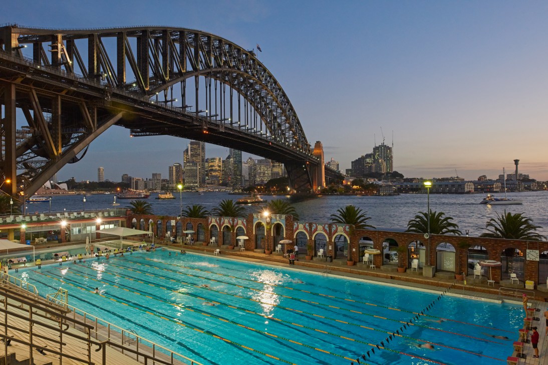 North Sydney Olympic Pool at dusk with Harbour Bridge and illuminated city skyline in background.