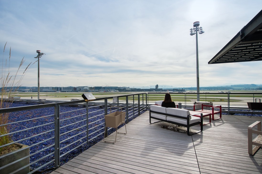 Woman seated on outdoor terrace at Swiss E Gate lounge overlooking airport runway with mountains in distance.