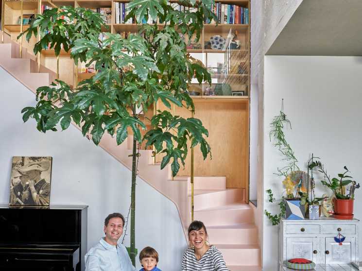 Family of three seated at wooden dining table in bright interior with pink staircase, bookshelves, and potted plant behind them.