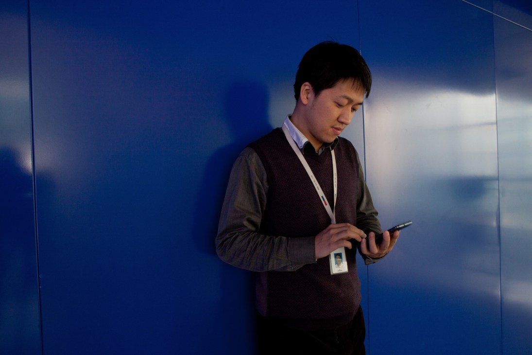 A young man in business attire holding a mobile device while standing in front of a blue wall.