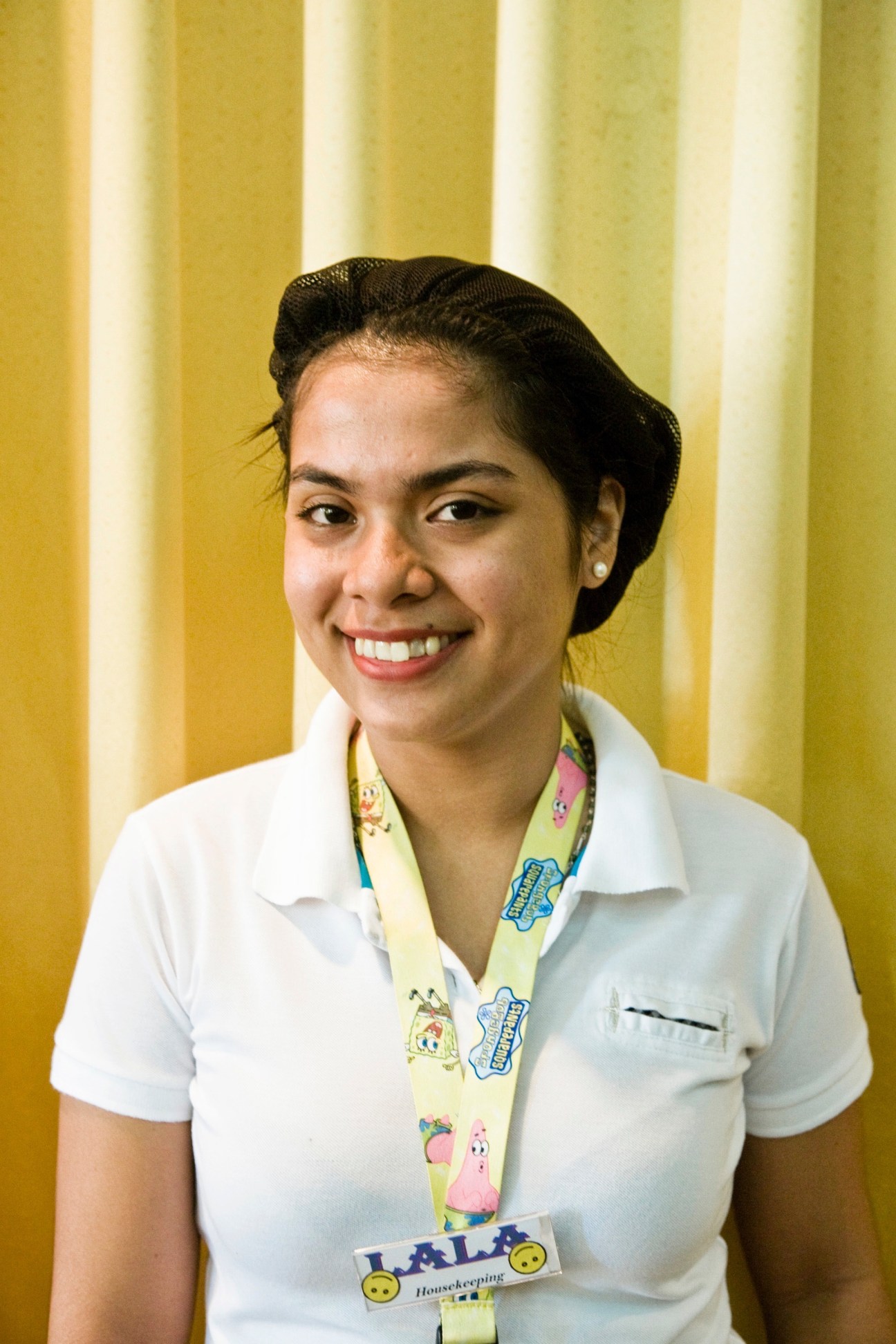 Young Filipino woman in white uniform smiling at camera with housekeeping identification badge.