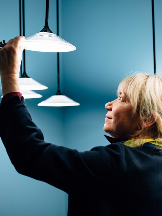 Woman examining a white pendant light fixture with black stem against blue wall.