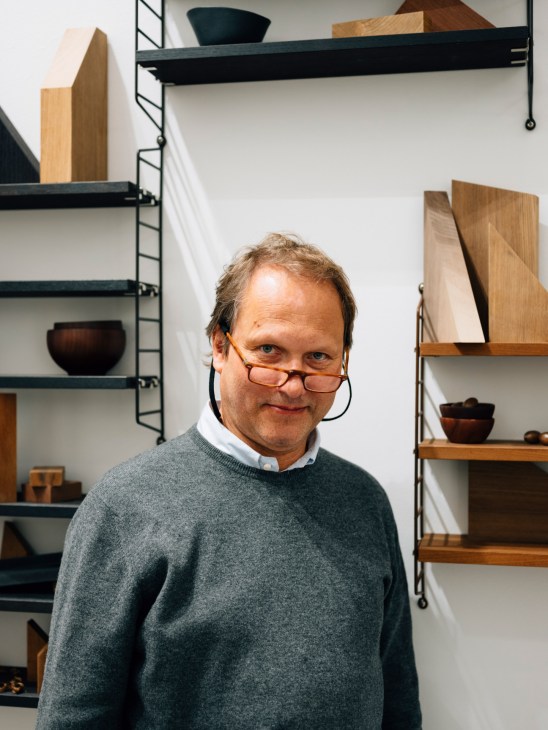 Peter Erlandsson, owner of String furniture, stands in front of minimalist shelving units displaying wooden objects and decorative items.