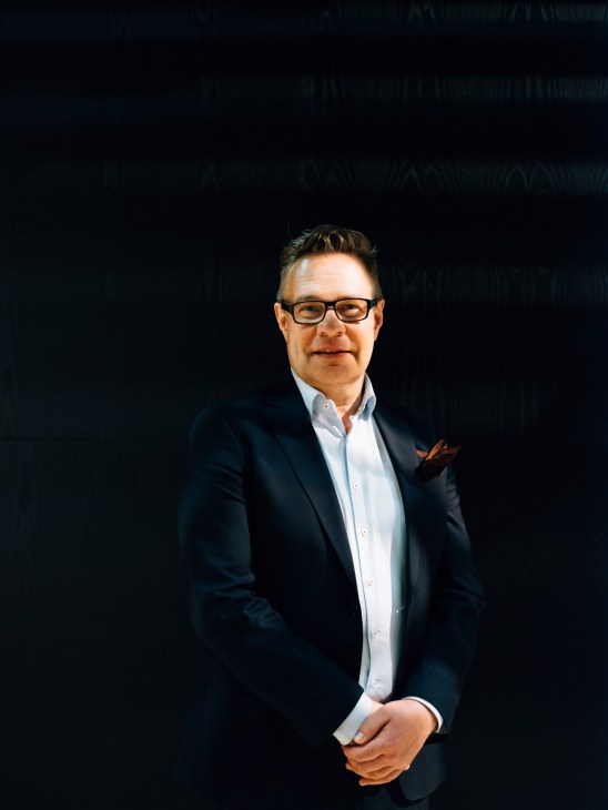Man wearing glasses and dark blazer with light blue shirt against dark background.