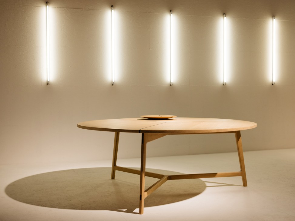 Modern wooden oval table with angular legs displayed under linear ceiling lights in a minimalist gallery space