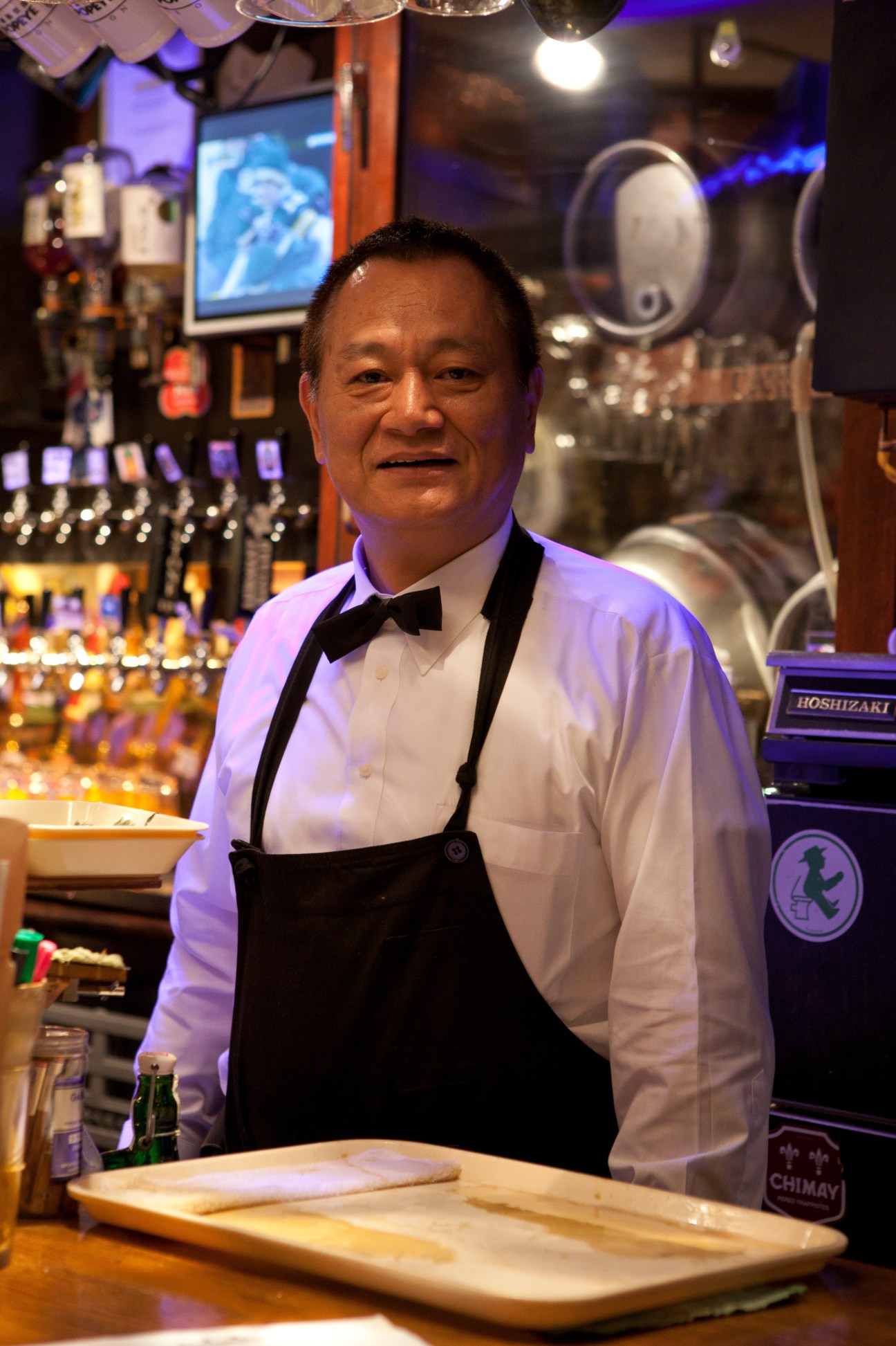 Bartender Tatsuo Aoki stands behind the bar at Popeye, wearing a white shirt and black apron.