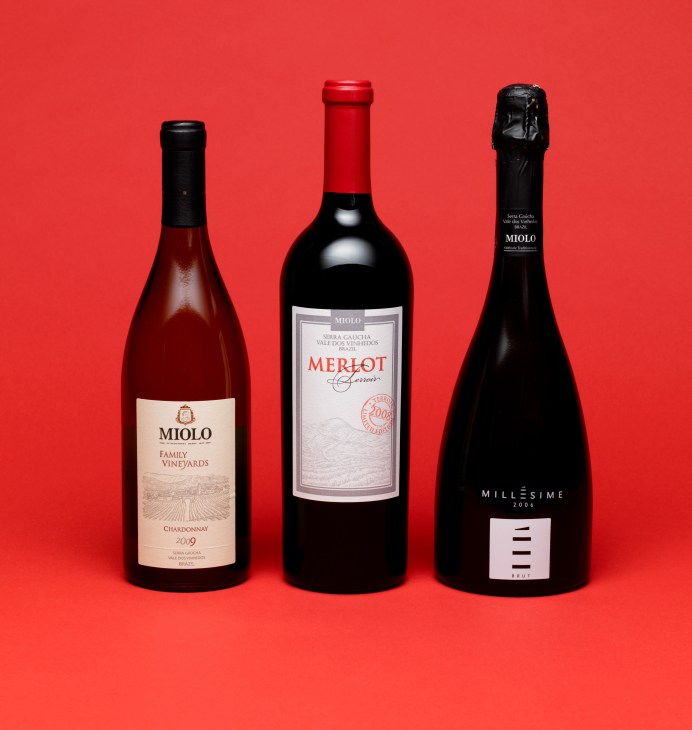Three Miolo wine bottles displayed against a red background: a Chardonnay, a Merlot, and a sparkling wine.