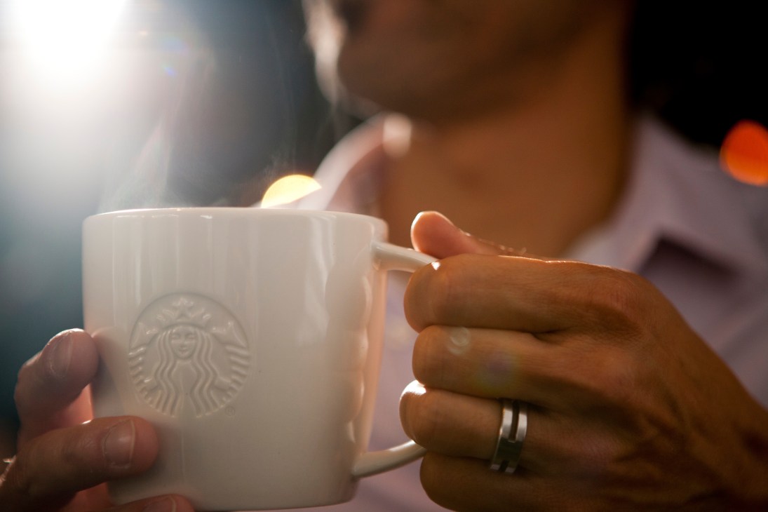 Person holding a white Starbucks mug with the siren logo in natural daylight.