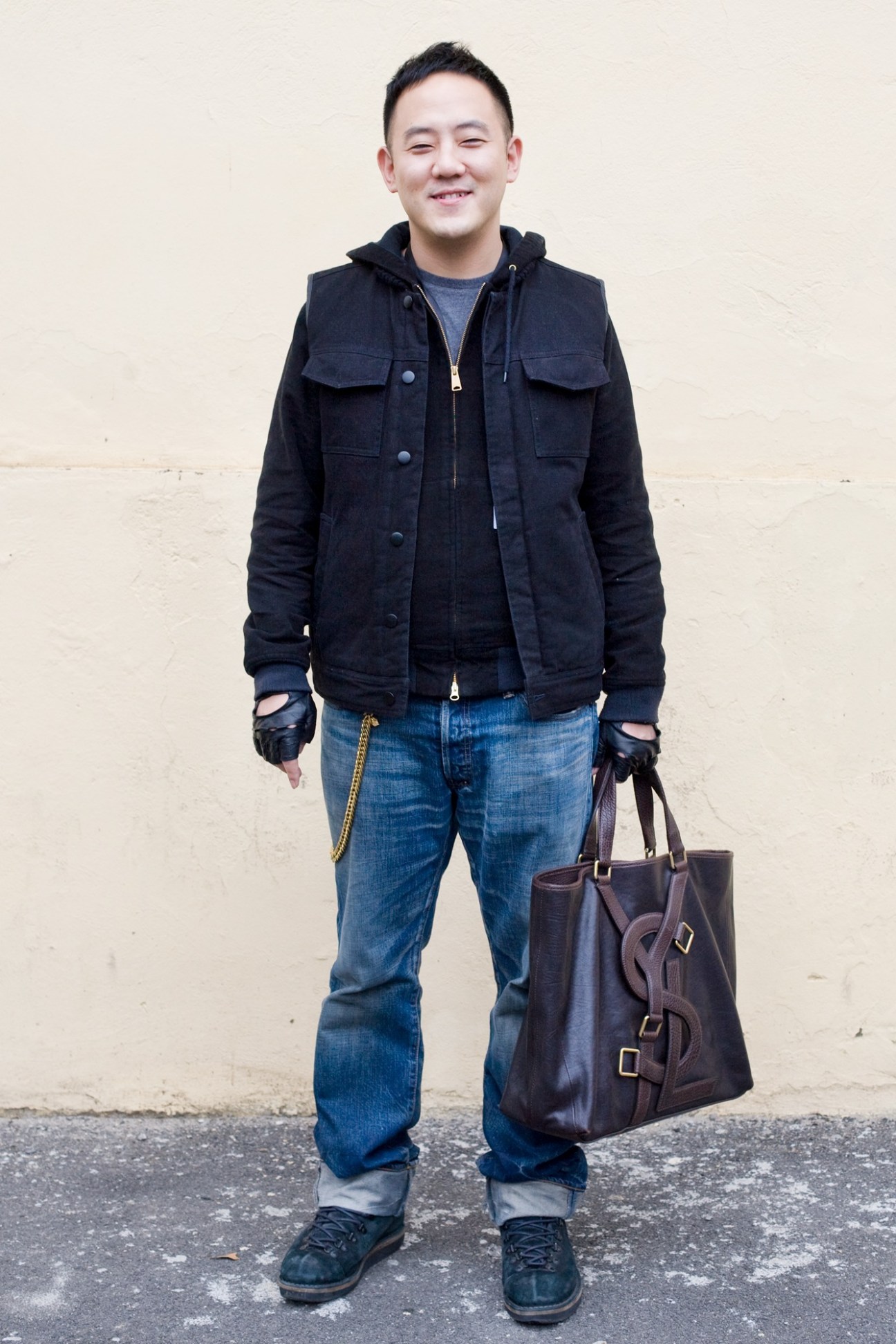 Man in dark denim jacket and blue jeans holding a brown leather bag.