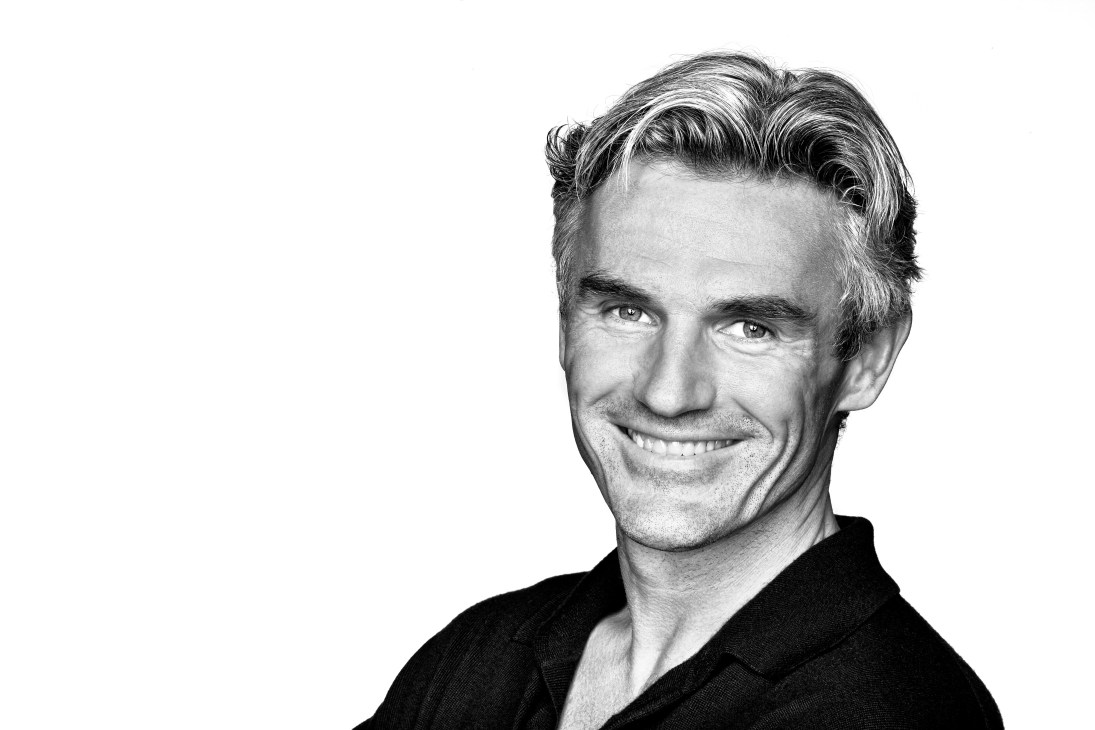 Black and white portrait photograph of a smiling man with styled hair wearing a black shirt.