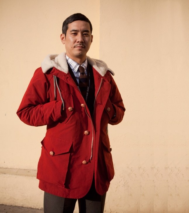 Man wearing red jacket with white collar, checked shirt and burgundy cardigan against beige wall.