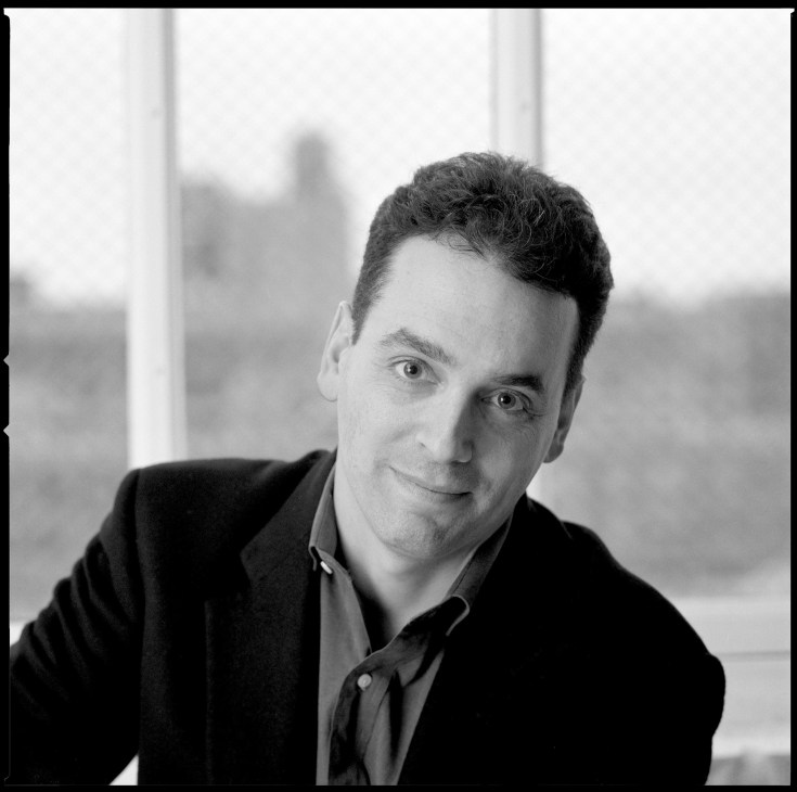 Black and white portrait photograph of Daniel Pink, author, by a window with blurred cityscape background.