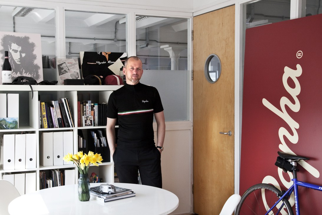 Simon Mottram, founder of Rapha cycling brand, standing in his office with the Rapha logo visible on the wall behind him.