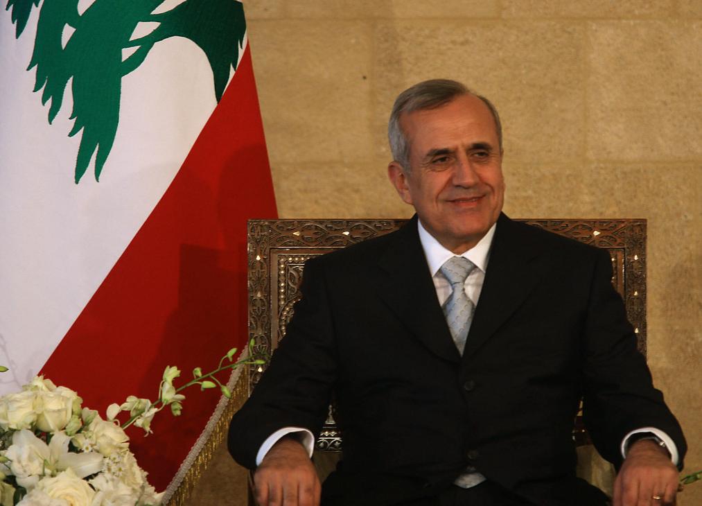 Man in formal black suit seated before Lebanese flag with white flowers.