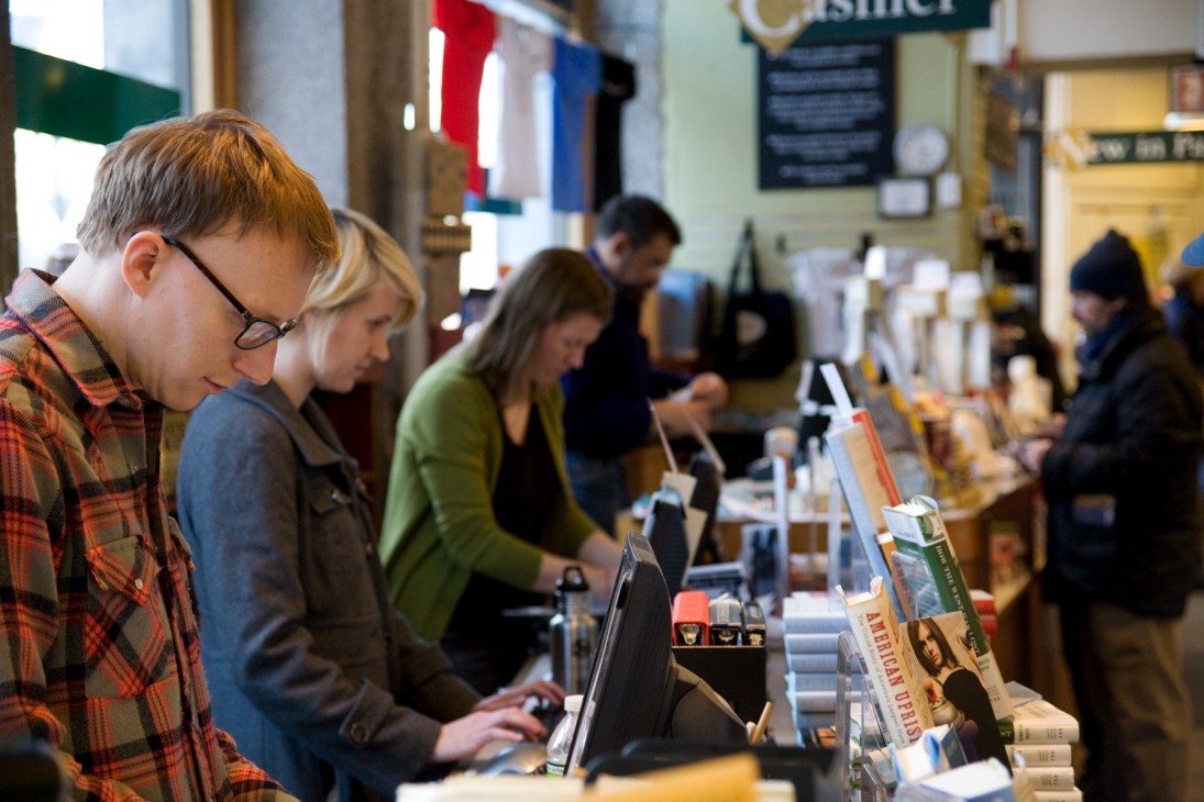Customers browse books at Harvard Book Store counter with staff assisting.