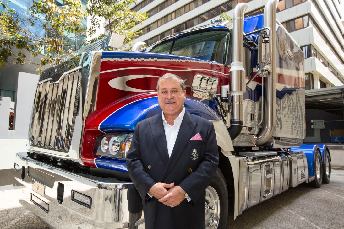 Man in dark blazer and pink pocket square stands in front of red and blue Mack truck with gold trim