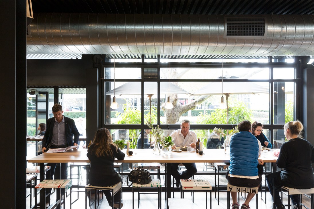 Interior of Dreieck Park bar in Fukuoka showing wooden counter, customers seated at tables near large windows overlooking a park.