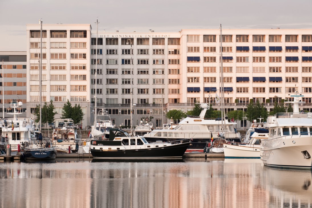 Waterfront view of Antwerp's harbor with moored boats and historic residential buildings reflected in calm water.