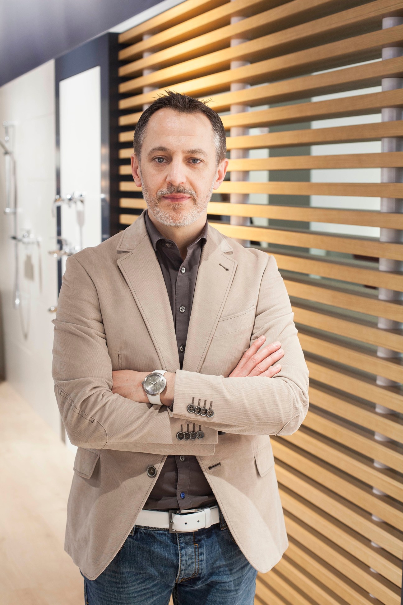 Man in beige blazer standing with arms crossed in front of wooden slat wall.
