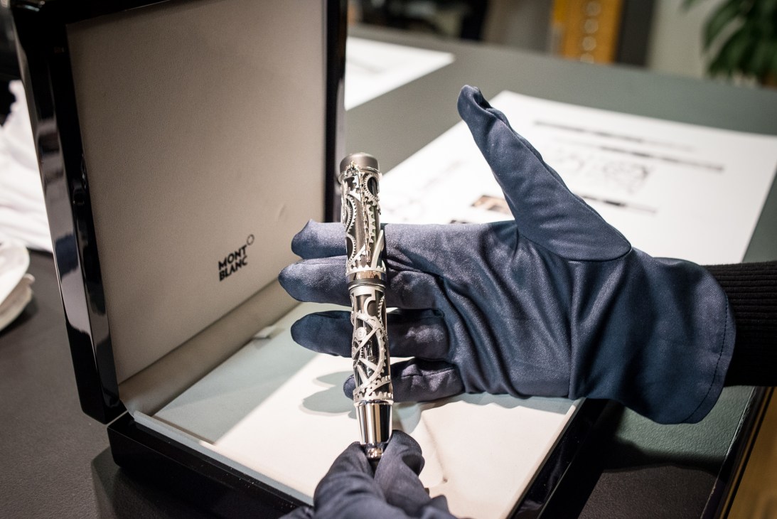 A Montblanc fountain pen with ornate silver details held in a gloved hand above blank paper.