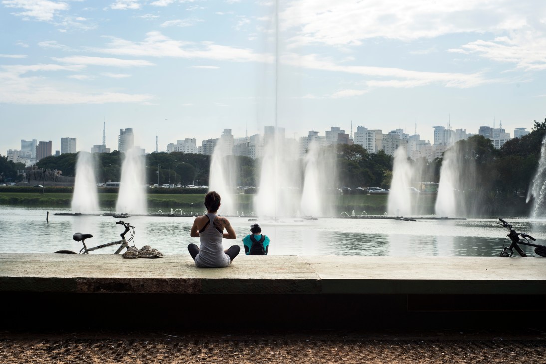 Woman and child sitting on sandy beach by lake with multiple fountains and city skyline in background.