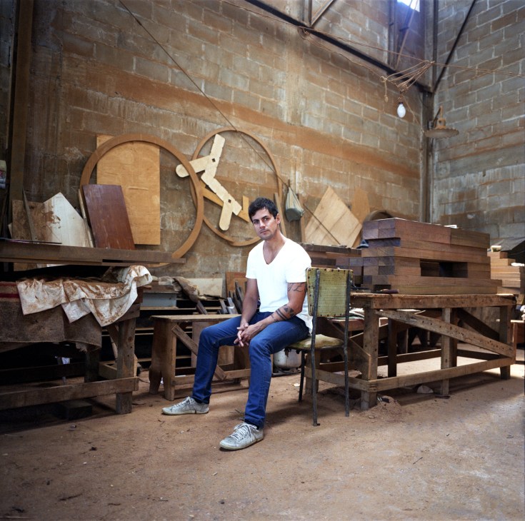 Zanini de Zanine sits in his Rio de Janeiro workshop surrounded by wooden furniture designs and sculptural pieces.