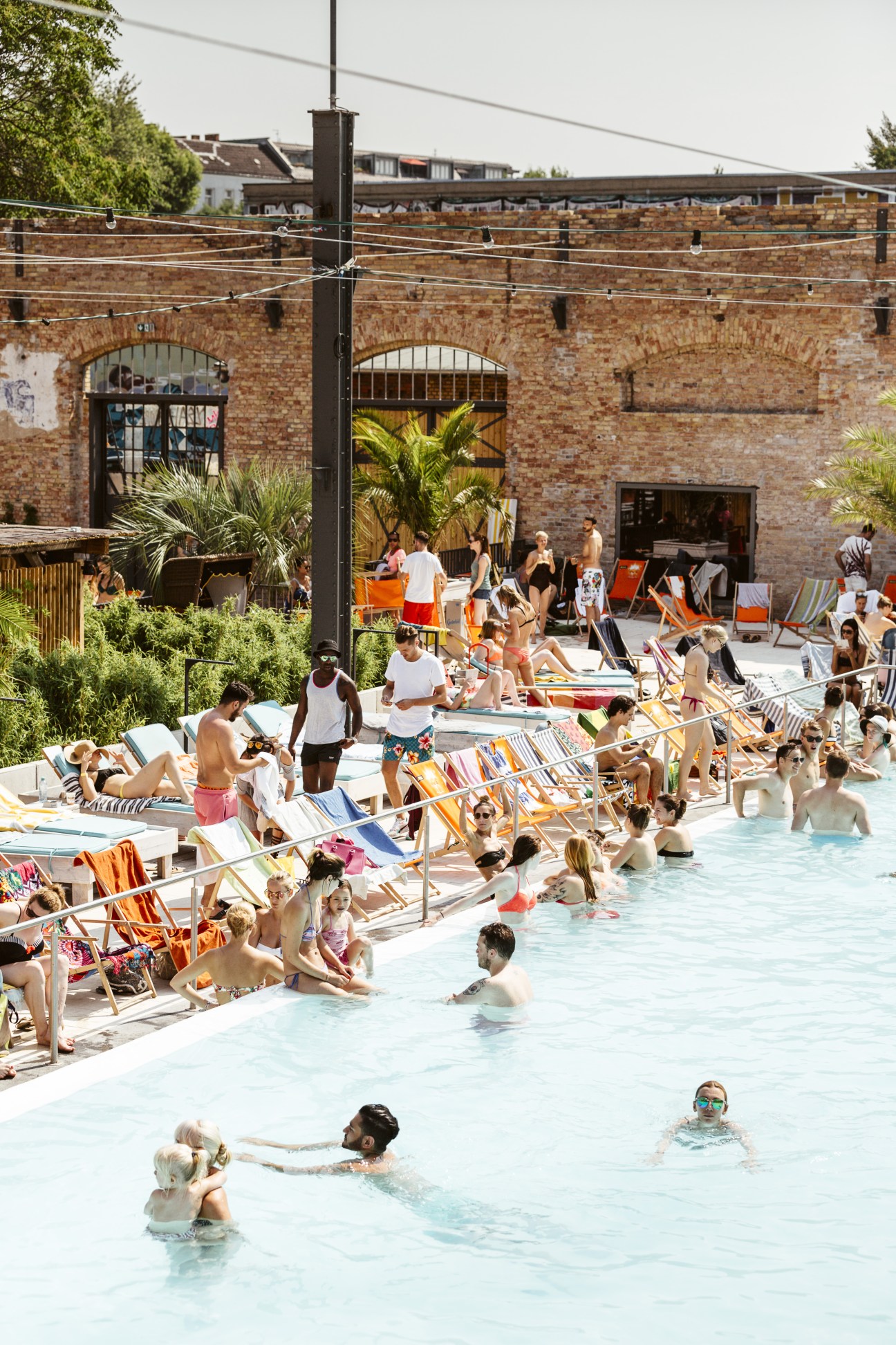 Haubentaucher pool with swimmers and sunbathers in colorful deck chairs against red brick industrial building.