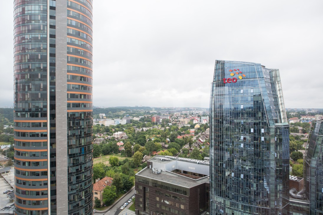 Modern high-rise buildings with orange and glass facades overlook a sprawling cityscape with green parks and residential areas under cloudy skies.