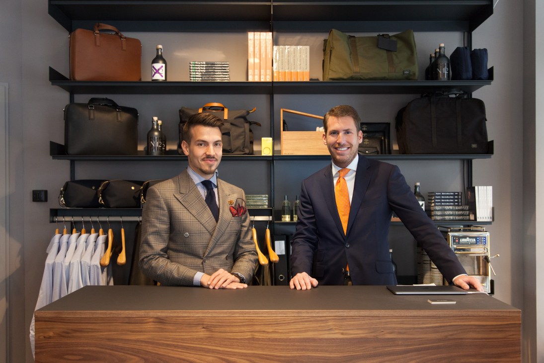 Two men in tailored suits stand behind a wooden counter in a menswear shop with shelves displaying clothing, accessories and bottles.
