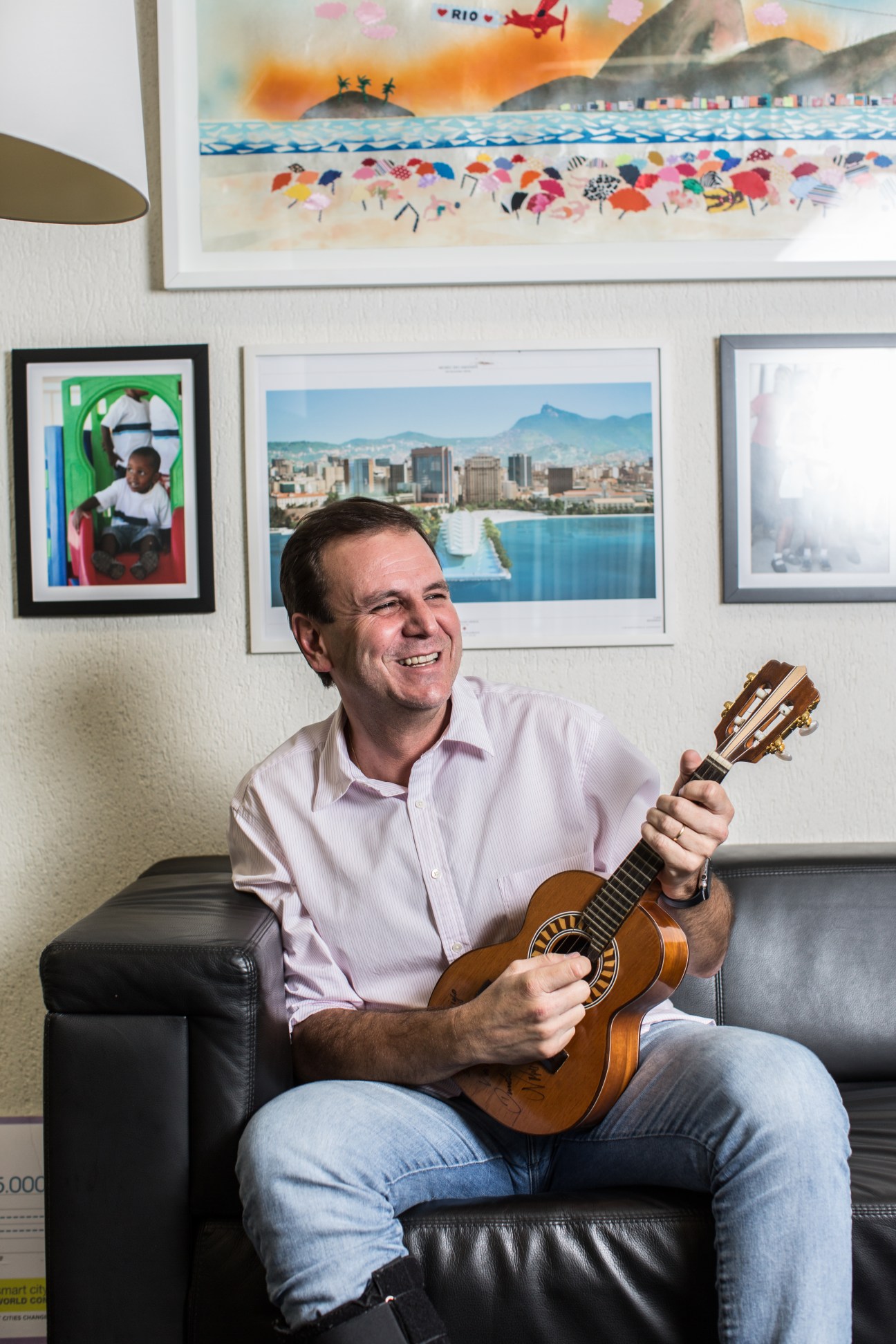 Man in pink shirt holding acoustic guitar, seated in office with Rio de Janeiro cityscape photo on wall.