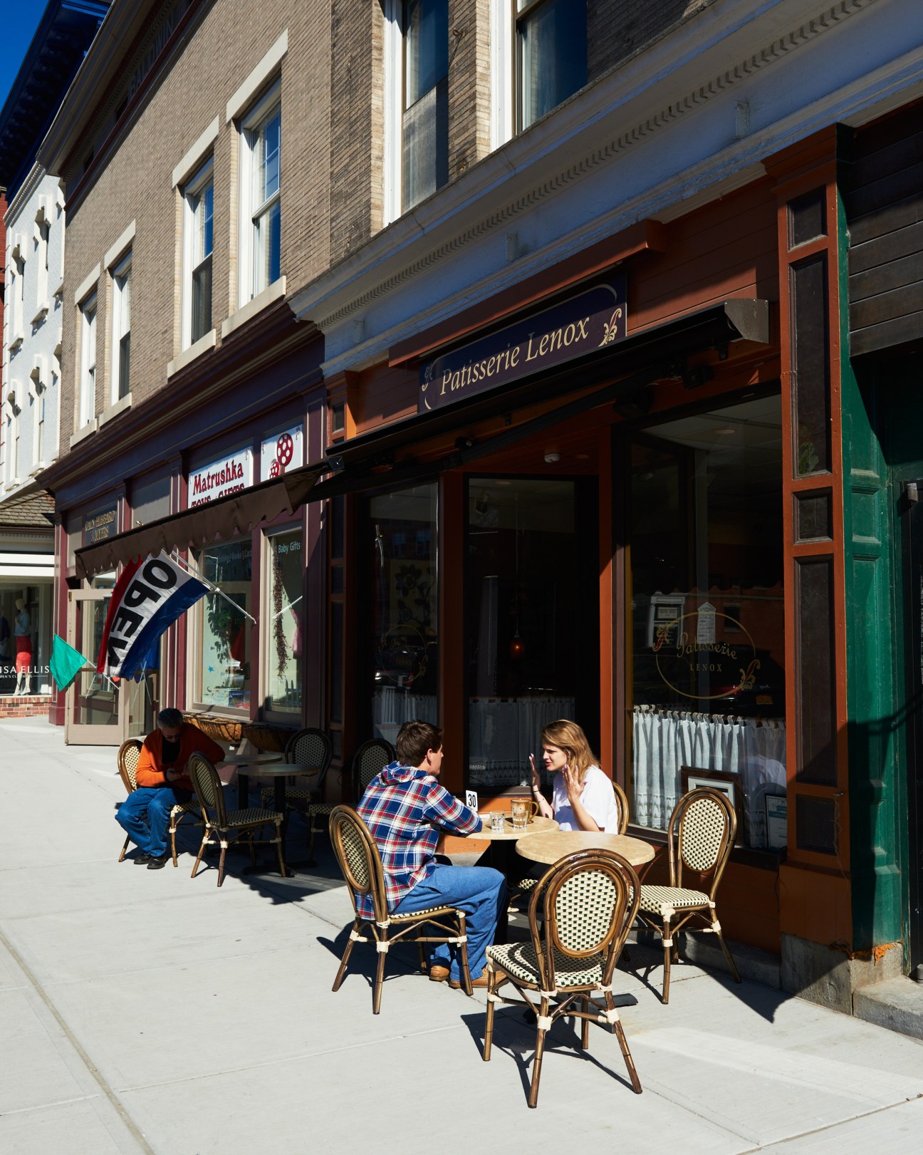People seated at outdoor café tables in front of storefronts on a sunny street.
